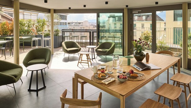 A modern dining area with a wooden table, green chairs, and a view of the city through large windows. - Base Geneva (Geneva)