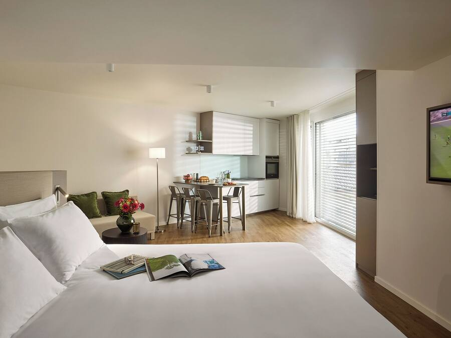 Una habitación de hotel con una cama grande, un escritorio pequeño, kitchenette y un ventanal amplio.