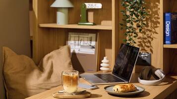 A wooden desk with a laptop, a glass of iced coffee, a plate with a pastry, a pair of headphones, and a small plant.