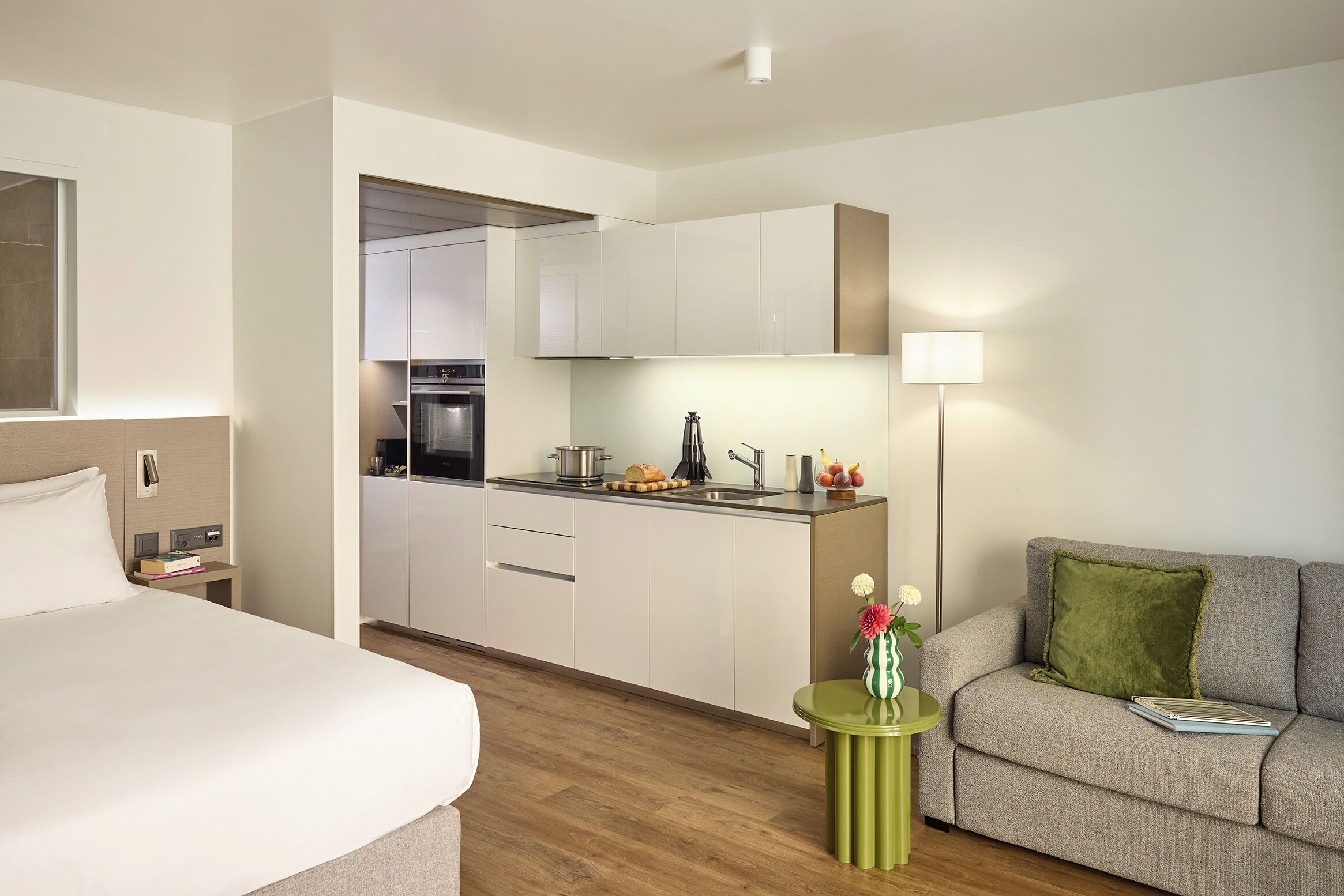 A modern hotel room with a bed, a green side table with a vase, a grey sofa with a green pillow, and a kitchenette with a stove and sink.