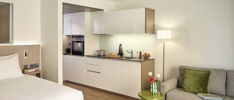 A modern hotel room with a bed, a green side table with a vase, a grey sofa with a green pillow, and a kitchenette with a stove and sink.