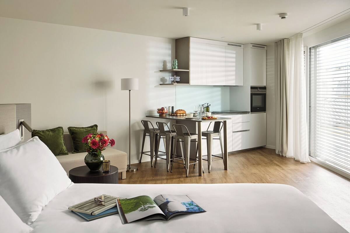 Une chambre d’hôtel moderne comprenant un lit, un coin lecture avec un magazine, une petite table agrémentée d’un vase de fleurs, une kitchenette et une grande fenêtre avec des stores.