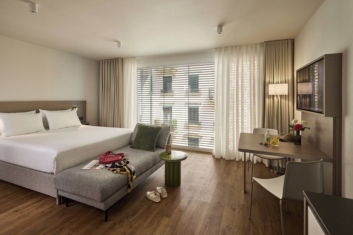 Une chambre d’hôtel avec un lit, une petite table avec une lampe, une télévision et une fenêtre avec des stores.