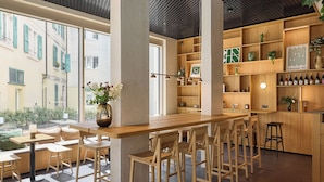 A modern bar area with wooden furniture, a large window with a view of buildings, and shelves with bottles and decorative items. - Base Geneva (Geneva)
