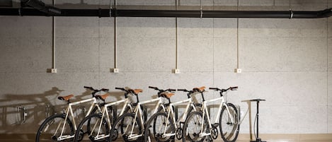 Varias bicicletas estacionadas junto a una pared en una habitación con piso y techo de concreto, y un patinete eléctrico a un lado.
