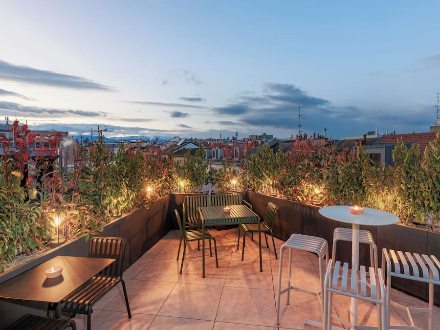 Een terras op het dak met een tafel en stoelen, omringd door planten en verlichting, met uitzicht op een stadsbeeld.