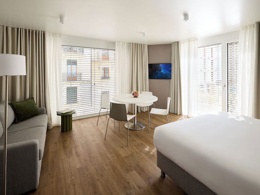A modern hotel room with a grey sofa, a round dining table with chairs, a bed, and a TV on the wall.