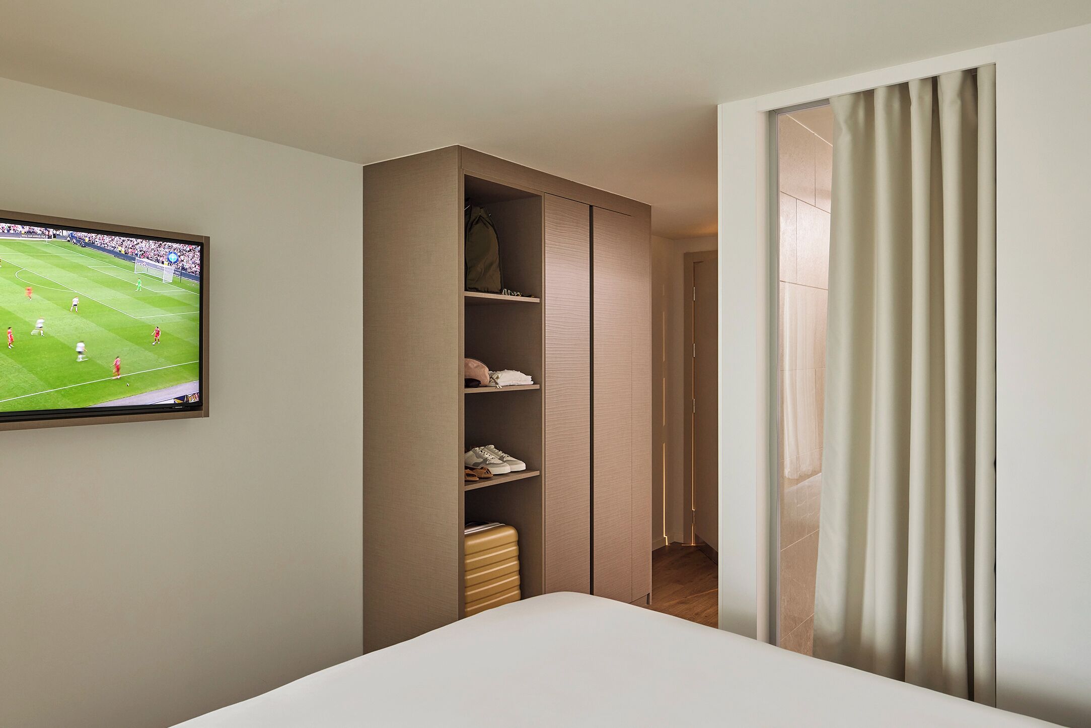 Una habitación de hotel con cama, armario, televisor mostrando un partido de fútbol y una cortina.