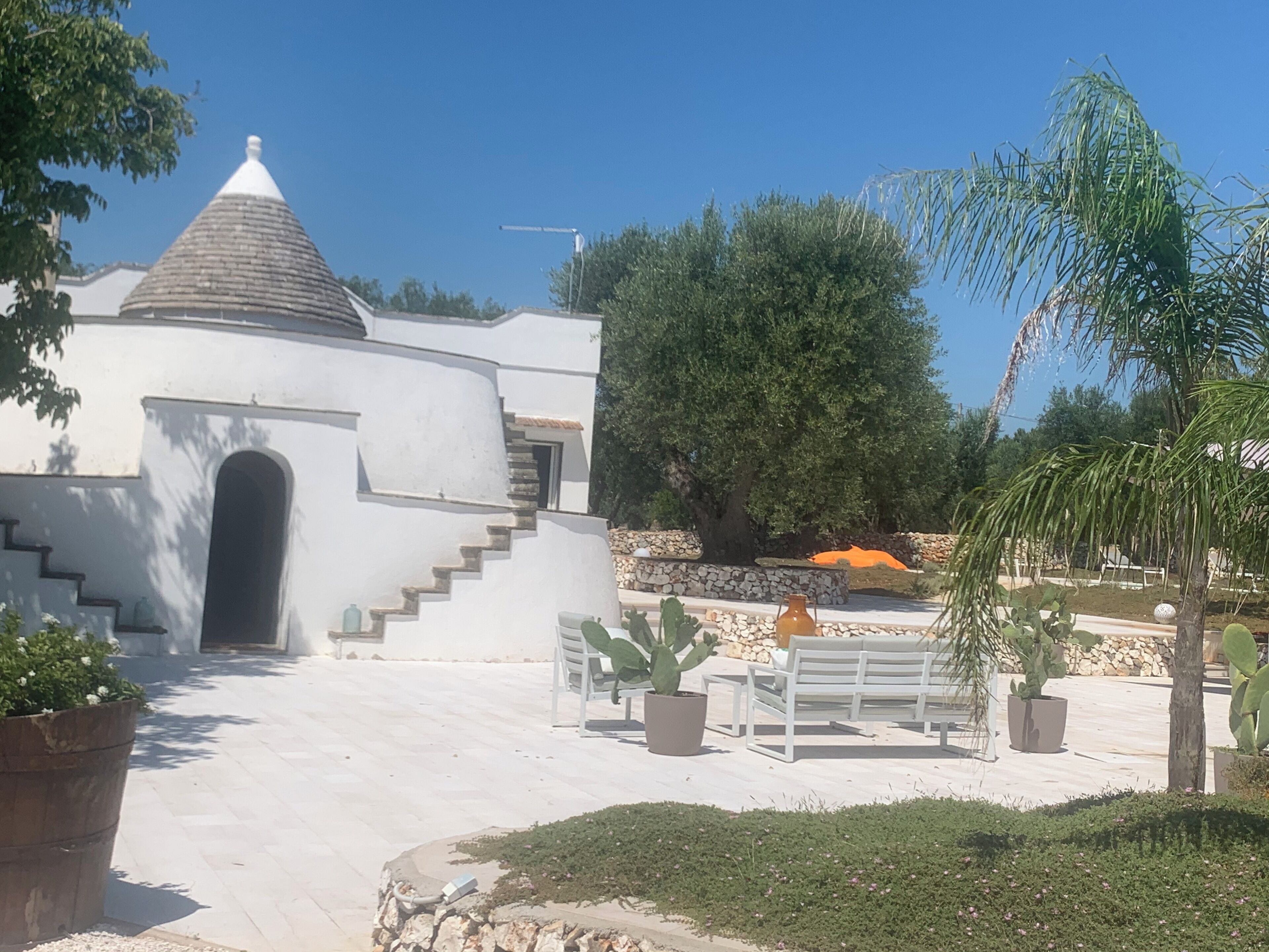Un bâtiment blanc coiffé d’un toit conique, doté d’un escalier en pierre et d’une cour dallée avec des bancs et des cactus.