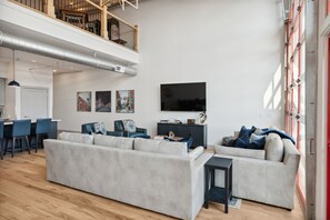 A spacious living area with a large sofa, a dining table, and a TV. There is a staircase leading to an upper level. - Majestic Lofts #601 - Penthouse (Nashville)