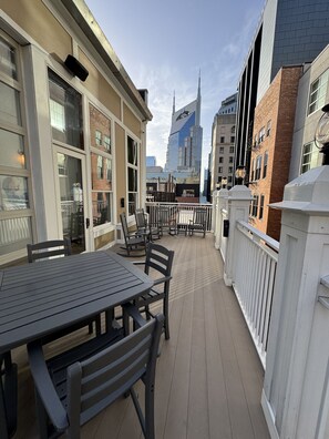A covered outdoor patio with a table and chairs, a white fence, and a view of a tall building in the background.