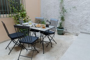 A small outdoor dining area with a table set for four, surrounded by chairs. There is a potted plant to the right and a window with a metal grill on the left. - 2BR Greek Home | Private Backyard 20' to Acropolis (Athens)