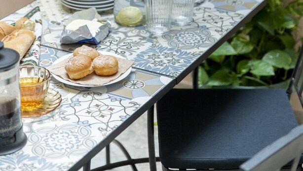 A table set for a meal with plates, glasses, and a pitcher, surrounded by green plants and a metal railing. - 2BR Greek Home | Private Backyard 20' to Acropolis (Athens)