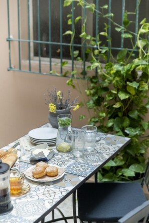 Mesa lista para una comida, con platos, vasos y una jarrón, rodeada de plantas verdes y una barandilla metálica.