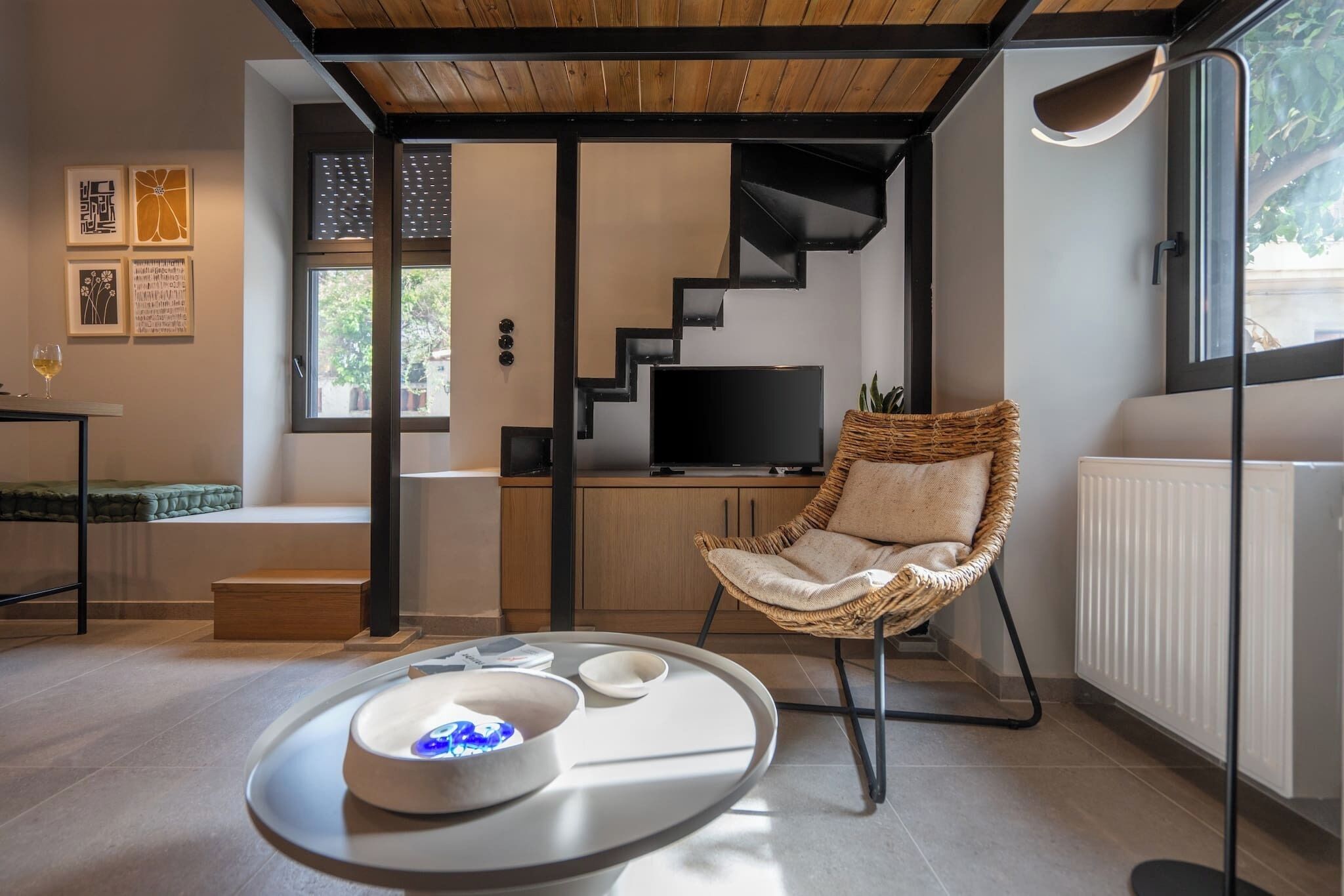Un salon moderne avec un plafond en bois, un escalier noir, un fauteuil en osier, un téléviseur à écran plat et une table à manger avec un verre de vin.
