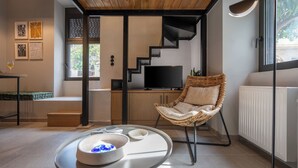 A modern living room with a wooden ceiling, a black staircase, a wicker chair, a flat-screen TV, and a dining table with a glass of wine. - 1BR Boutique Apt Acropolis Rooftop view (Athens)