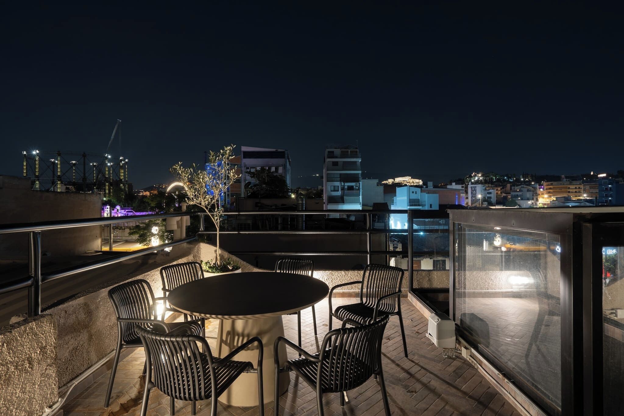 A rooftop terrace with a round table and chairs, overlooking a cityscape at night.