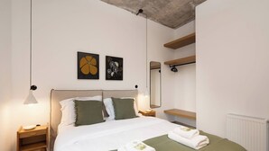 A neatly made bed with two pillows, a green throw blanket, and two folded towels on the bed. There is a bedside table with a lamp, a wall-mounted mirror, and two framed artworks above the bed. - 2BR Suite Apt • Acropolis Rooftop Views • By Metro (Athens)