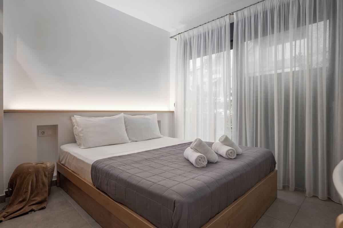A modern hotel room with a large bed, white curtains, and a wooden headboard.