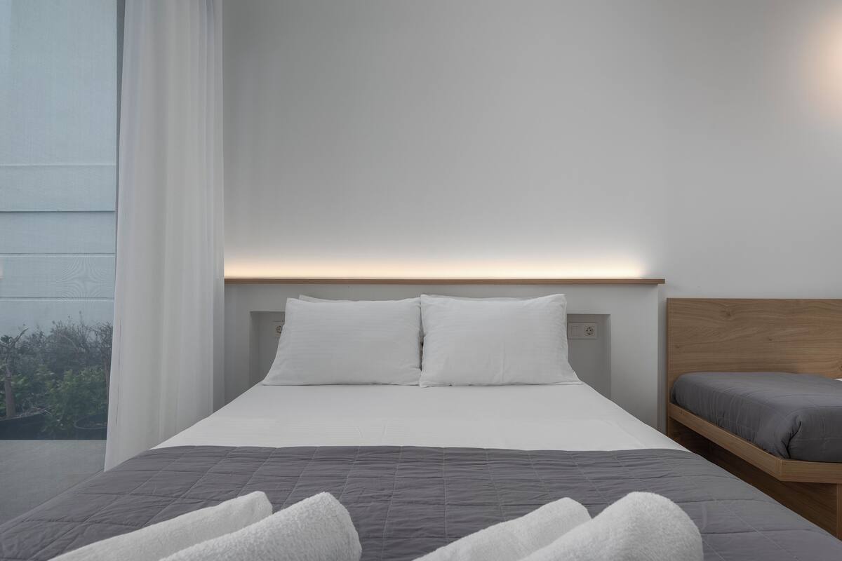 A modern hotel room with a large bed, a smaller bed, and a view of greenery outside.