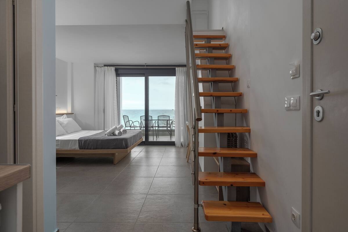 A modern bedroom with a bed, a wooden ladder, a balcony with a view of the sea, and a door with a handle.