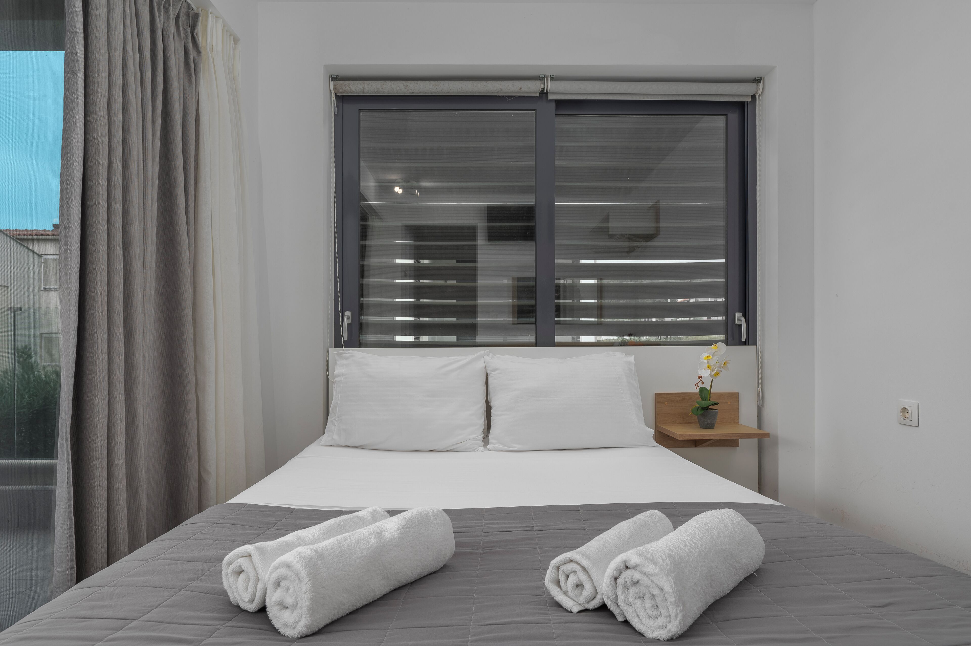 A bedroom with a bed, two pillows, a window with blinds, a small wooden shelf with a plant, and two rolled towels on the bed.