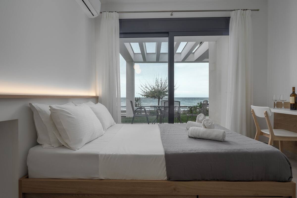 A modern bedroom with a large bed, a view of the sea, and a small table with a bottle and glasses.