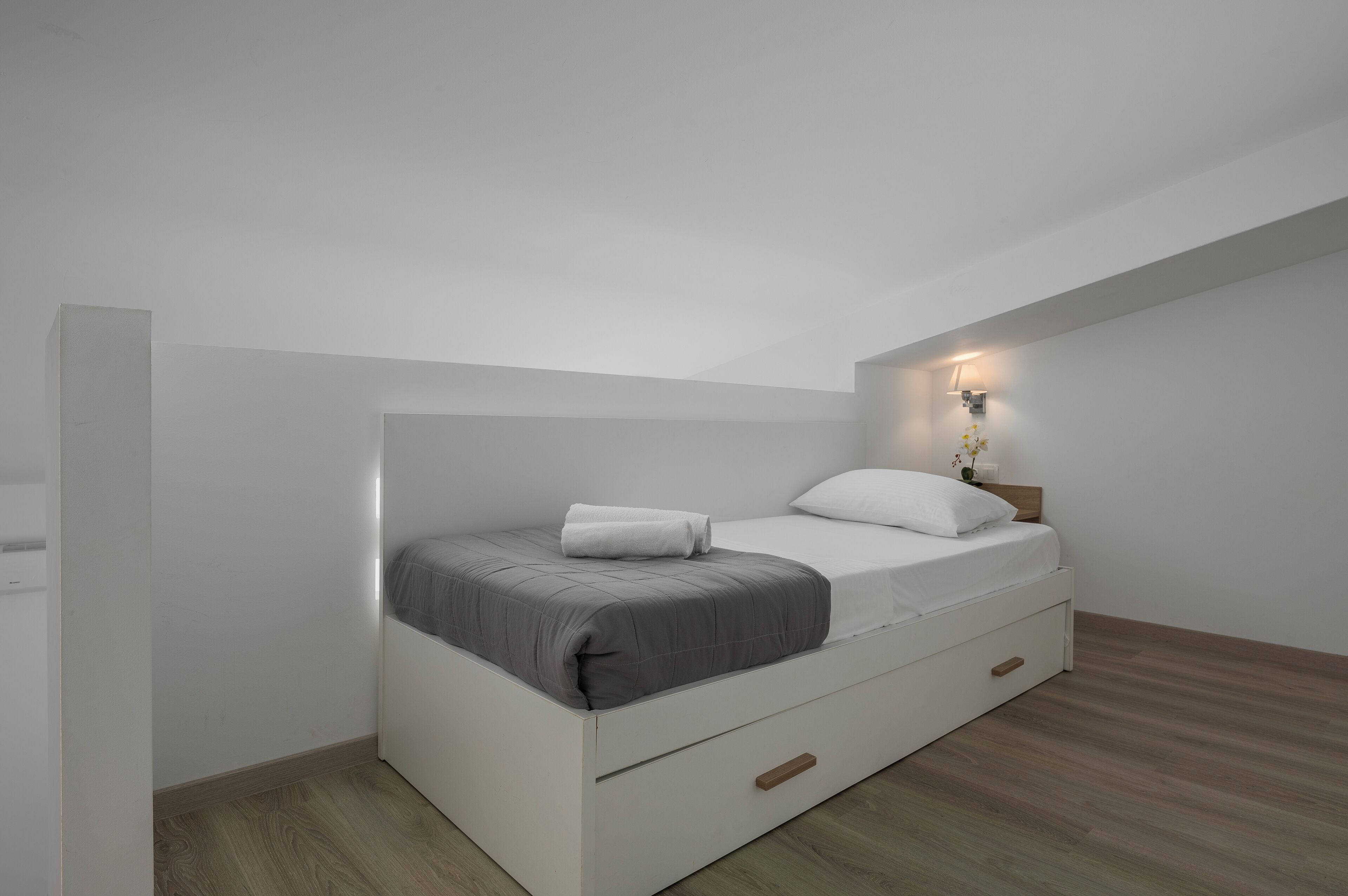 A modern, minimalist bedroom with a single bed, a nightstand, and a wall-mounted light fixture.