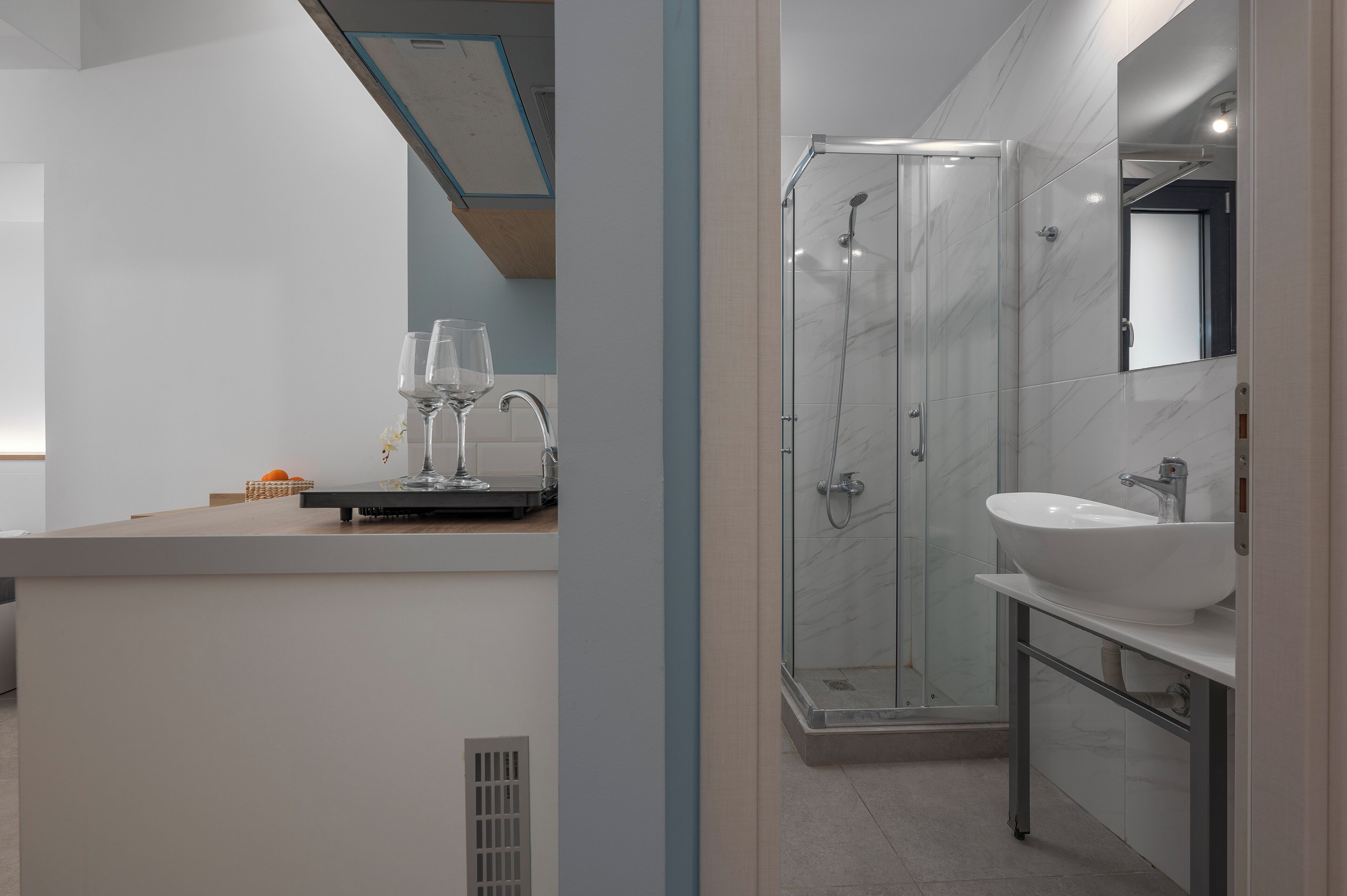 A modern bathroom with a glass-enclosed shower, a white sink, and a countertop with a wine glass and oranges.