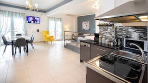 A modern kitchen with a black stove, a built-in sink, and a microwave. There is a dining area with a table and chairs, and a yellow armchair. A TV is mounted on the wall, and a chandelier hangs from the ceiling.