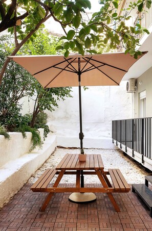 Una mesa de picnic de madera con una sombrilla color beige, rodeada por un patio de ladrillo y follaje.