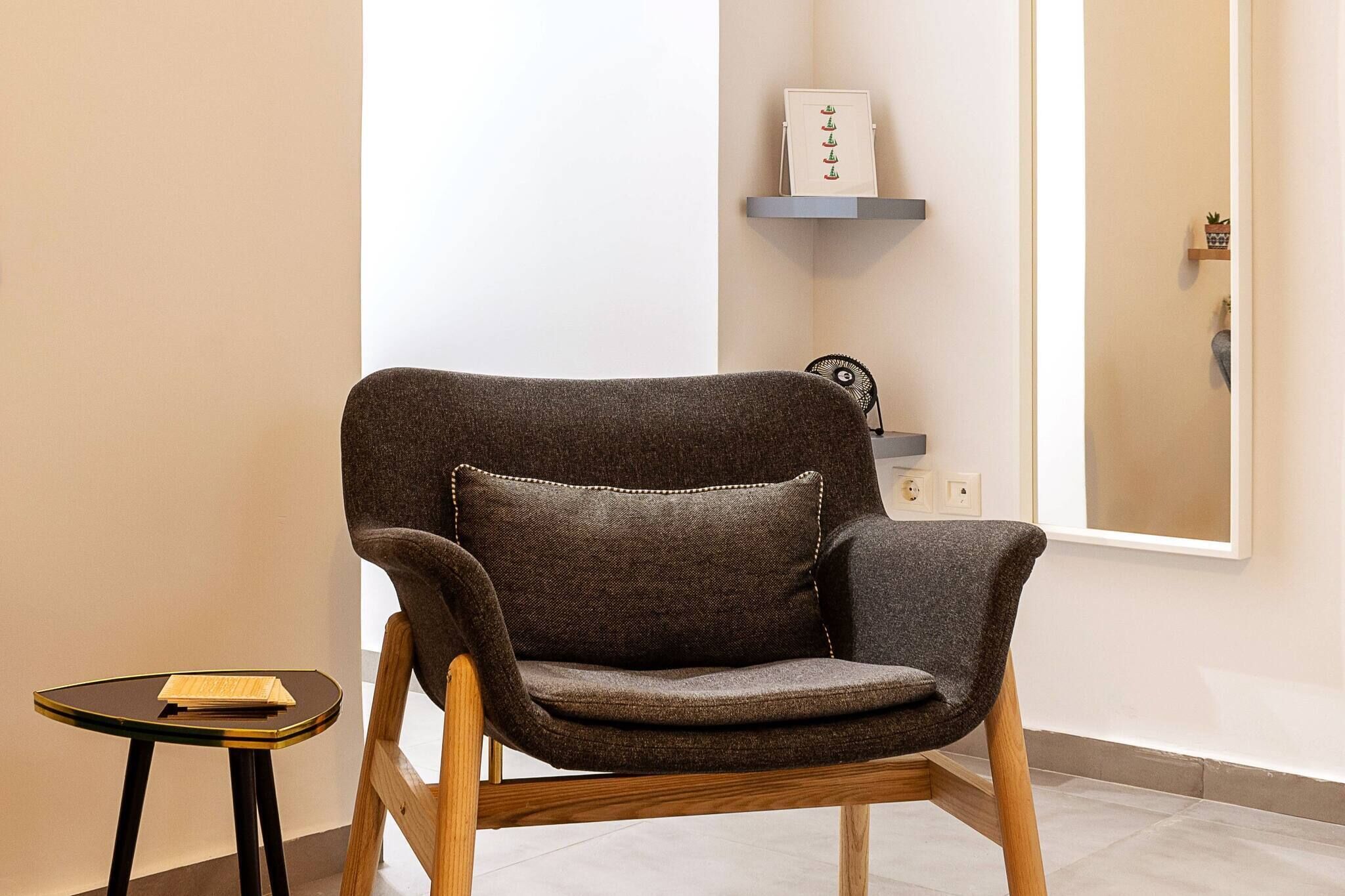Een moderne fauteuil met kussen, een bijzettafeltje met een boek, en een wandplank met decoratieve objecten.