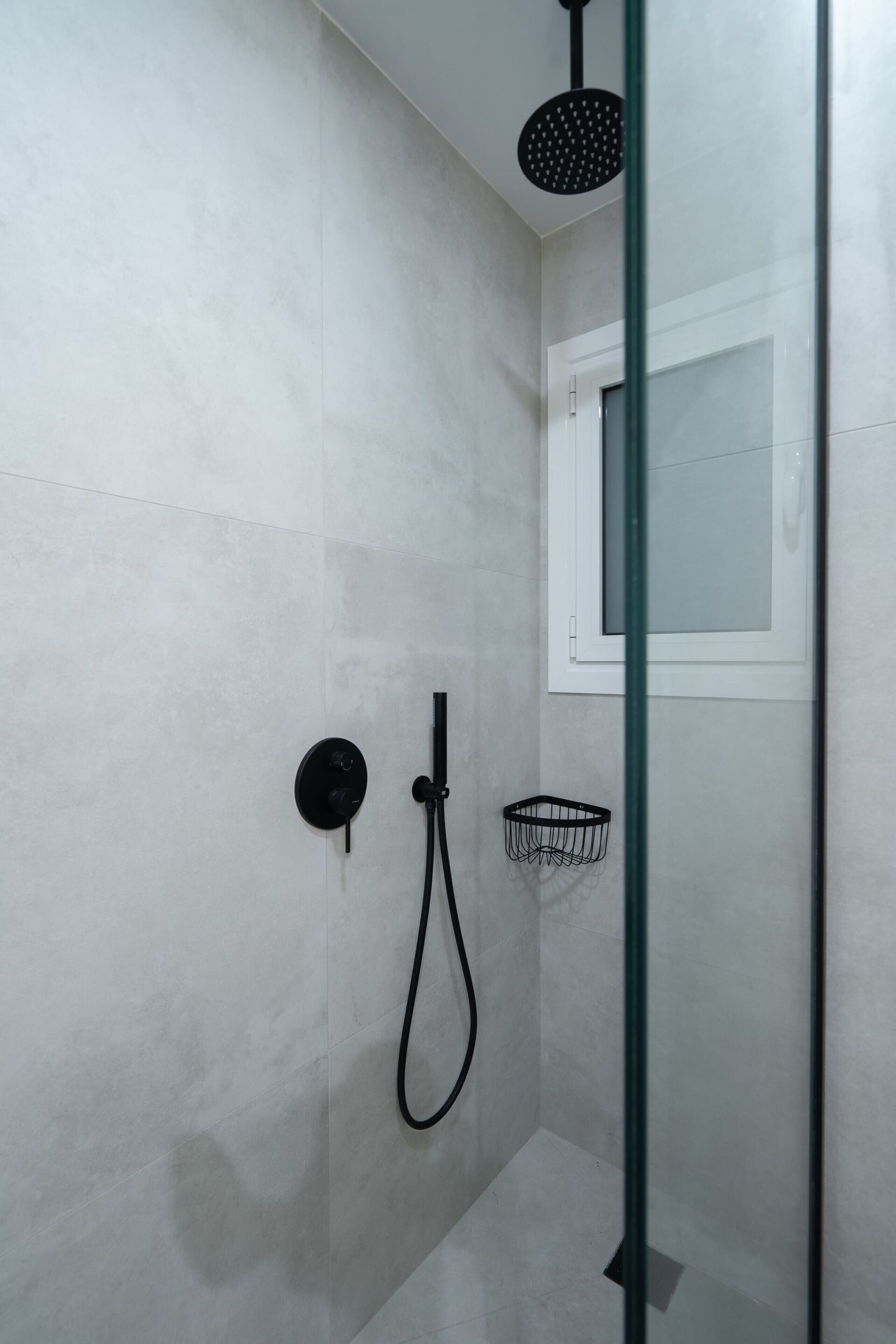 Une douche équipée d’un pommeau de douche à main, d’un panneau de commande mural et d’un petit panier noir.