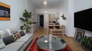 A modern living room with a sofa, a glass coffee table, a dining area with chairs, a kitchen in the background, and a TV mounted on the wall. - Urban Escape: Exarcheia Remodel (Athens)