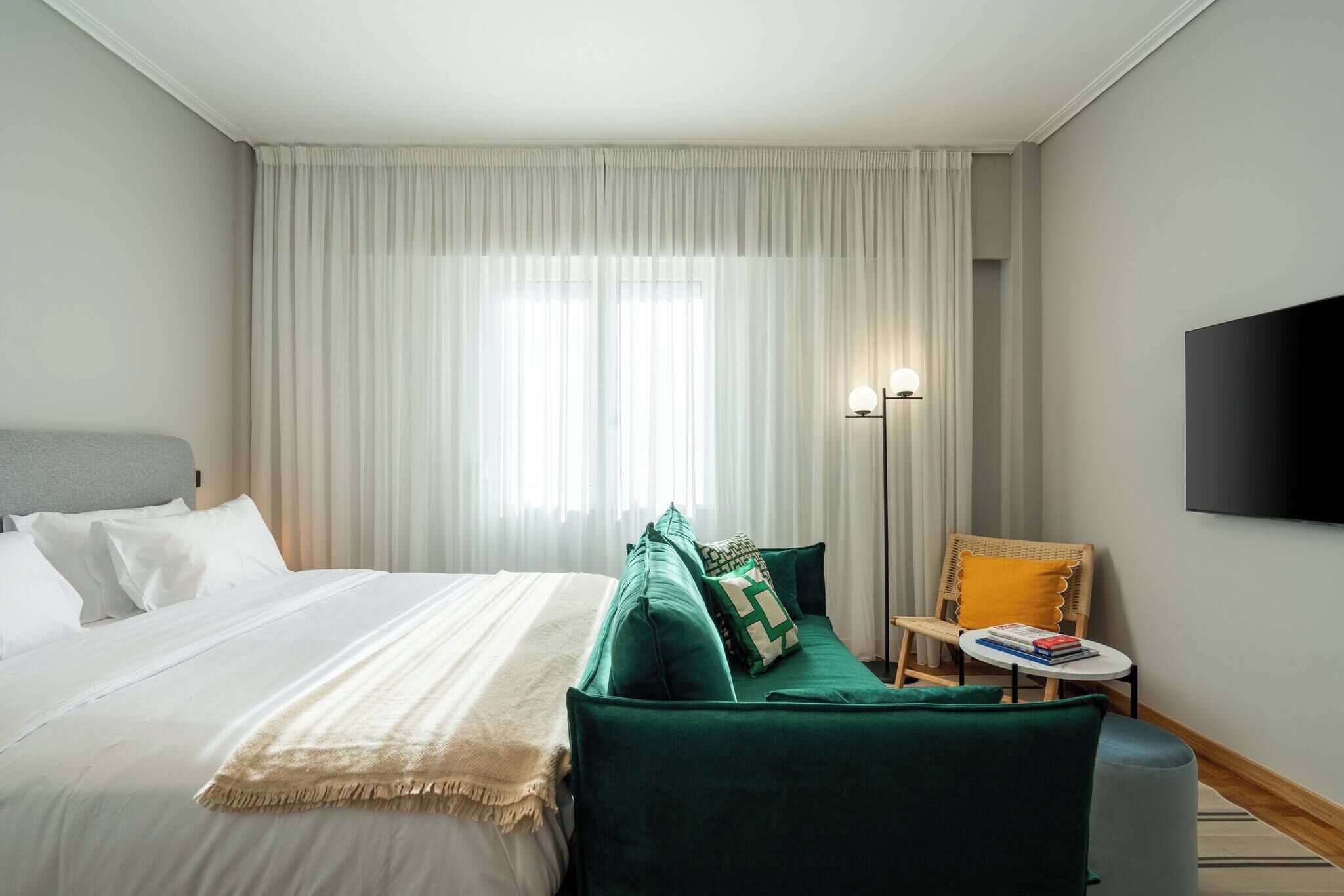 Hotellihuone, jossa on sänky, vihreä sohva, pieni pöytä, jossa on kirjoja, lamppu ja seinälle asennettu televisio.