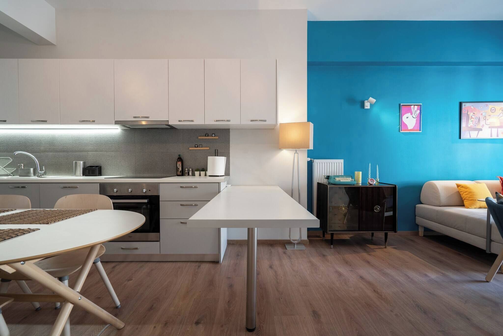 Een moderne keuken met witte kastjes, een eettafel en een bank in een woonruimte met een blauwe accentmuur.