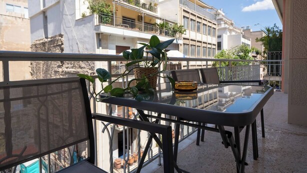 A balcony with a glass table and chairs, surrounded by a metal railing, overlooking a residential area with multi-story buildings. - Exclusive Apt In The  Of Athens'_2BD+2 Bathrooms (Athens)