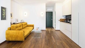 A modern living room with a yellow sofa, a glass coffee table, and a kitchen with white cabinets and a wooden countertop. - Athens' Best Location~ Unique Magnificent Studio (Athens)