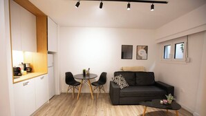 A modern living room with a black sofa, a round coffee table, and a dining table with chairs. There is a kitchen area with white cabinets and a wooden countertop. - 1BR In Plaka | Rooftop Access w/ Acropolis View (Athens)