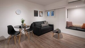 A modern living room with a black sofa, a round dining table with chairs, a small side table, and a wall clock. - 1BR In Plaka | Rooftop Access w/ Acropolis View (Athens)