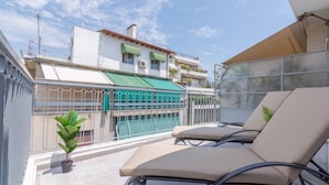 Un balcón con dos sillones de descanso, una pequeña planta en maceta, una piscina y un edificio con balcones al fondo.