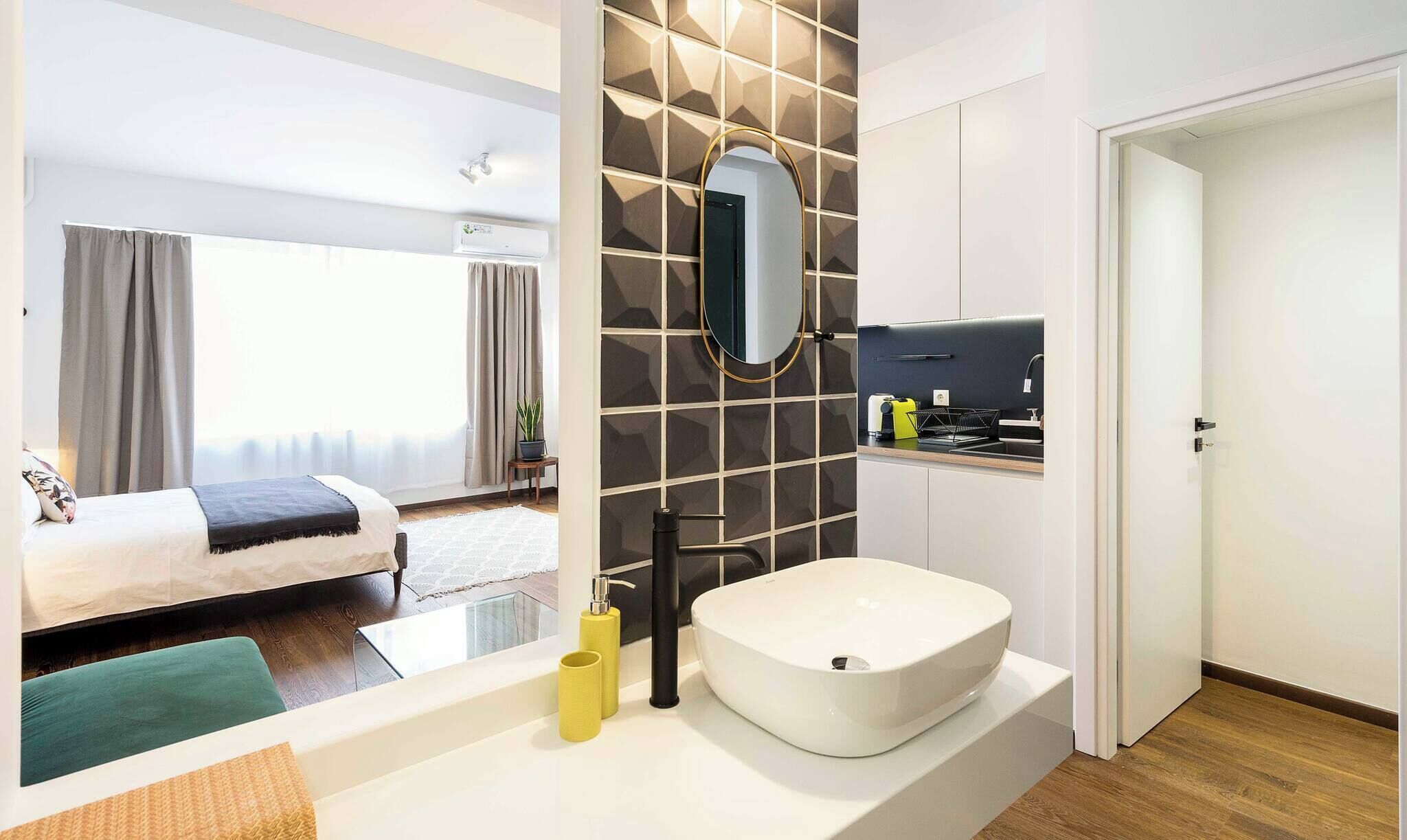 A modern hotel room with a large bed, a round white sink, a mirror, and a small kitchen area.