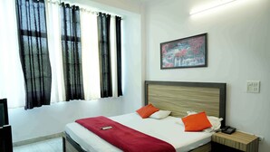 Un lit bien fait, recouvert d’une couverture rouge, avec deux oreillers orange et une tête de lit en bois. Un tableau représentant une cascade est accroché au mur, et un téléphone se trouve sur la table de chevet.