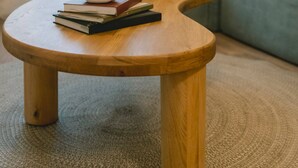 Une table basse ronde en bois, agrémentée de livres et d’un vase, est placée dans un salon où se trouvent un canapé gris et des coussins à motifs.