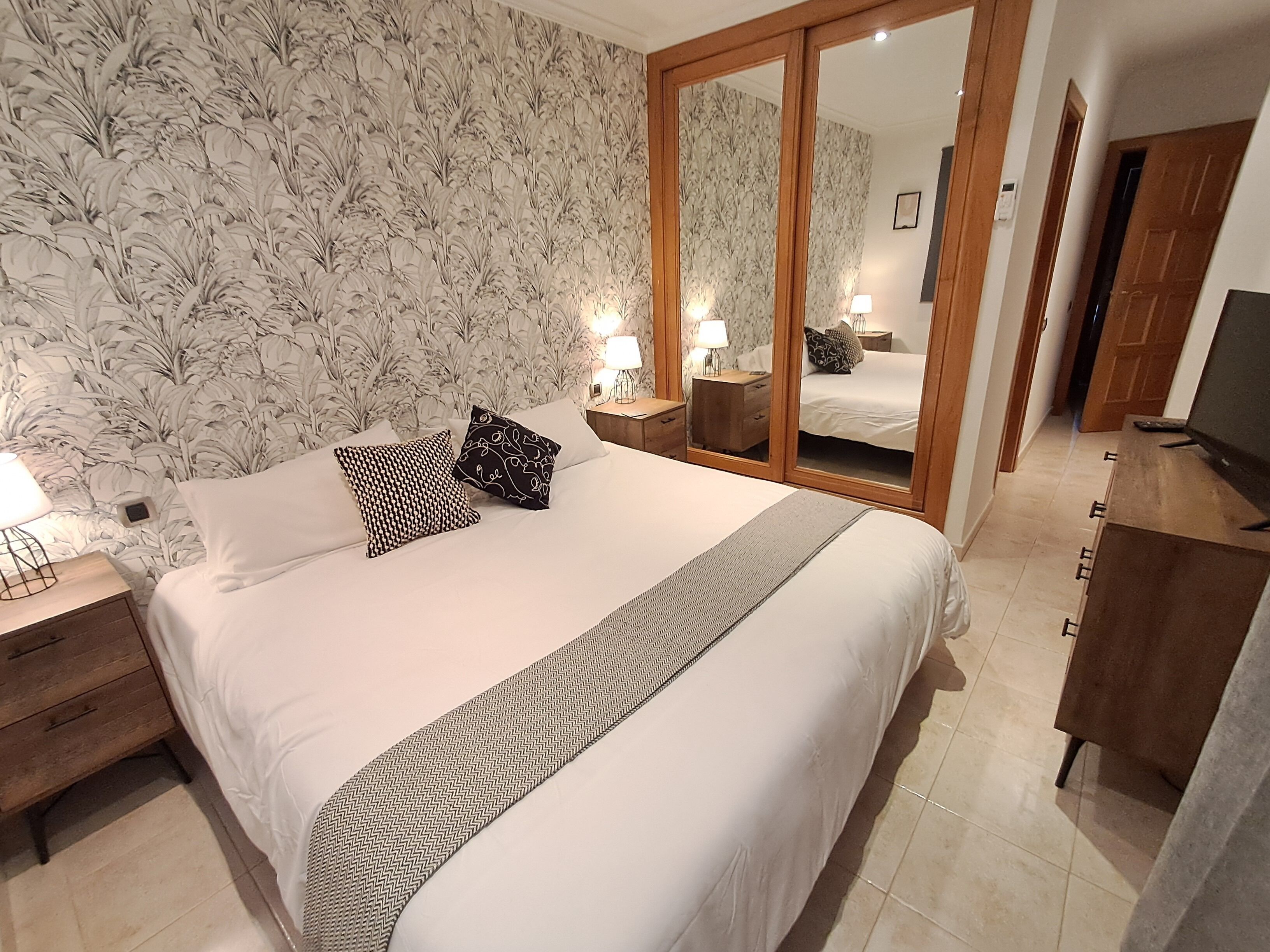 A neatly made bed with white linens and two patterned pillows, a wooden nightstand with a lamp, a large mirror, and a wooden dresser with a television.