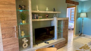 Una sala de estar con televisor, estanterías con libros y objetos decorativos, un mueble de madera y una lámpara.
