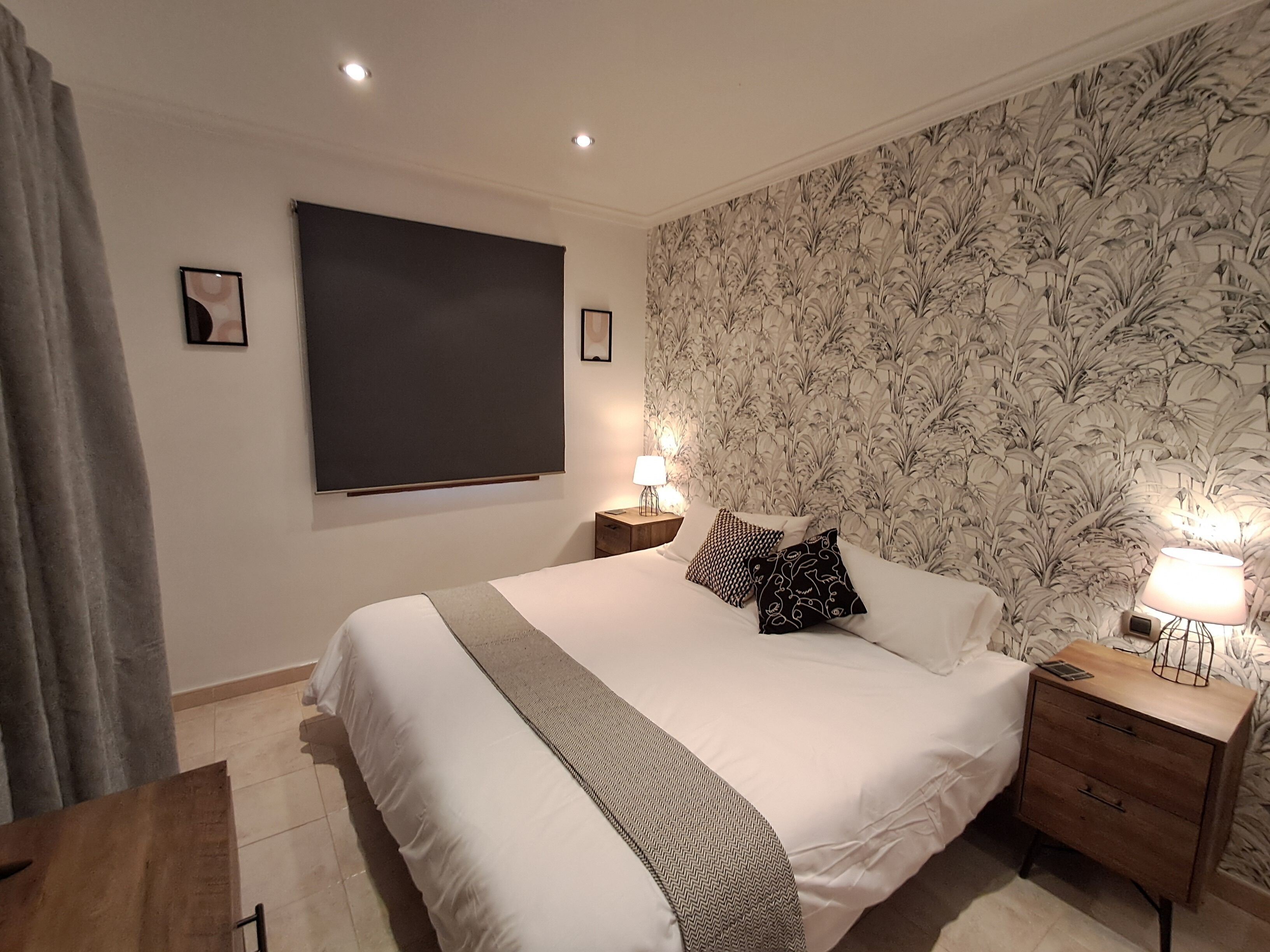A bedroom with a large bed, two bedside tables with lamps, a window with a blind, and a floral wallpaper.