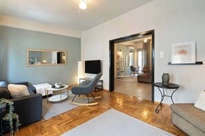 Un salon moderne avec un canapé, un fauteuil à bascule, un téléviseur et un coin repas visible à travers une porte ouverte.
