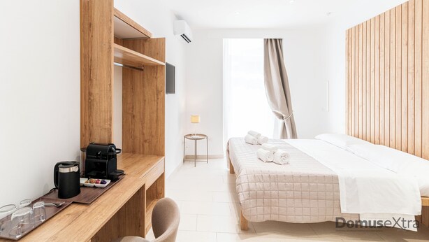 A modern hotel room with a wooden desk, a bed with a beige quilt, a small table with a lamp, and a view of the outdoors through a window with curtains. - Olivia's Home By DomusExtra (Naples)