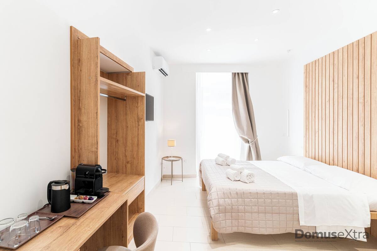 Camera d'albergo moderna con una scrivania in legno, un letto con copriletto color beige, un tavolino con lampada e vista sull'esterno attraverso una finestra con tende.